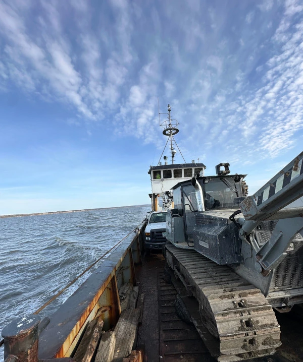 A large boat crossing the water, carrying a white pickup truck and a large gray tracked vehicle